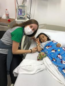 Jovem intercâmbista ao lado da cama de hostpital conversando com uma criança