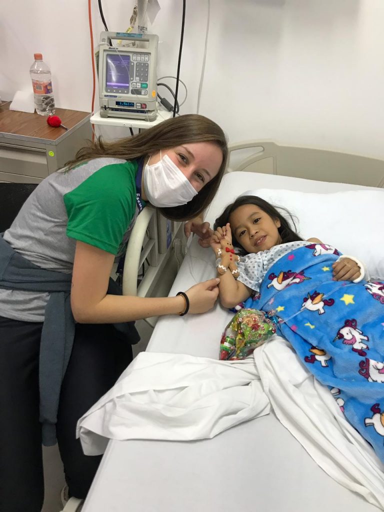 Jovem intercâmbista ao lado da cama de hostpital conversando com uma criança